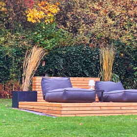 Gartenbereich mit einer modernen Holzdeck-Lounge, großen Polstermöbeln, umgeben von hohen Pflanzkübeln mit Gräsern und buntem Herbstlaubhintergrund