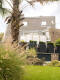 Gartenansicht mit modernen xxl Pflanzkübeln, gefüllt mit verschiedenen Gräserarten, vor einem Haus mit sichtbarer Terrasse und Freizeitmöbeln