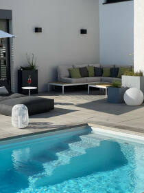 Moderne Poolterrasse mit Loungebereich, Gartenmöbeln mit Kissen, grauen Kübeln, einer weißen runden Designerlampe und einem erfrischenden Swimmingpool an einem sonnigen Tag