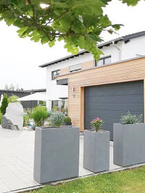 Einfahrt mit hohen Pflanzkübeln in grau vor einem Haus mit Holzverkleidung und grauem Garagentor