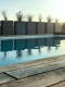 Abendstimmung am Pool mit einer Reihe hoher, rechteckiger Pflanzkübel, die sich im ruhigen Wasser spiegeln, Holzdeck im Vordergrund