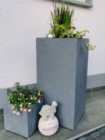 Eingang eines Hauses mit hohen und niedrigen grauen Pflanzkübeln, bepflanzt mit Ziergräsern und Blumen, neben einer dekorativen Schneckenfigur mit Willkommensschriftzug