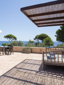 Terrasse mit Meerblick, ausgestattet mit Holzliege und Sonnenschutz aus Schilfrohr, umgeben von mediterraner Landschaft