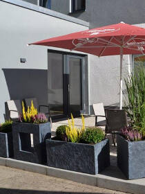 Außenbereich eines Cafés mit lava anthraziten rechteckigen Pflanzkübeln mit lebendigen Pflanzen und rotem Sonnenschirm