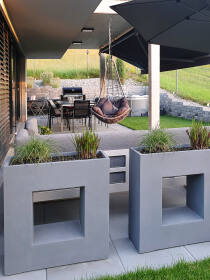 Moderne Terrassengestaltung mit grauen Vista Pflanzkübeln, integrierten Sitzgelegenheiten, Hängesessel, Grillbereich und Sonnenschirm, umgeben von einer gepflegten Rasenfläche und Hangbepflanzung