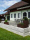 Ländliche Gartenterrasse mit Steinmauer, rustikalen Pflanzkübeln in rostfarbe, Gartenmöbeln mit Metallstühlen und Tischen vor weißen Haus mit Ziegeldach
