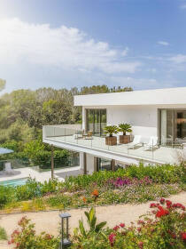 Moderne Luxusvilla mit weißer Fassade und großzügiger Terrasse, umgeben von einem farbenfrohen Garten mit Blick auf eine grüne Landschaft