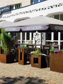 Außenbereich eines Cafés mit RatsHerrn Sonnenschirmen und rustikalen rost Pflanzkübeln mit bunten Blumen auf einem Kopfsteinpflasterplatz
