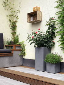 Ecke einer gemütlichen Terrasse mit modernen hohen und niedrigen Pflanzkübeln in Grau, bepflanzt mit Rosen und Kräutern, neben einer Lounge-Ecke, Holzboden und einer dekorativen Wandkiste