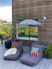 Terrassengestaltung mit lilanen Sitzsäcken, orangen gestreiften Kissen, einem schwarzen runden Pflanzenkübel mit Grünanlage und Sonnenschirm vor einer Holzfassade