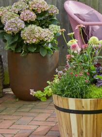 Gartenecke mit Blumen in hölzernen Fass-Pflanzgefäß und rostfarbenen runden Pflanzkübel mit Hortensien neben rosa Gartenstuhl