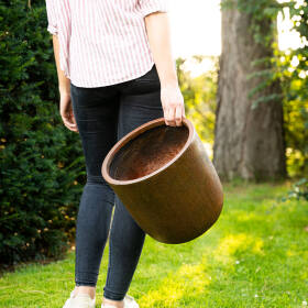 Frau läuft und hält einen leichten rostfarbenen Pflanzkübel rund im Garten