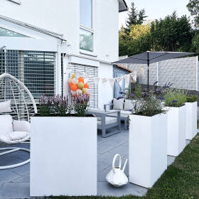Gartenterrasse mit weißen Pflanzkübeln groß, bepflanzt mit lila Blumen und Gräsern, neben Gartenmöbeln und Hängeschaukel